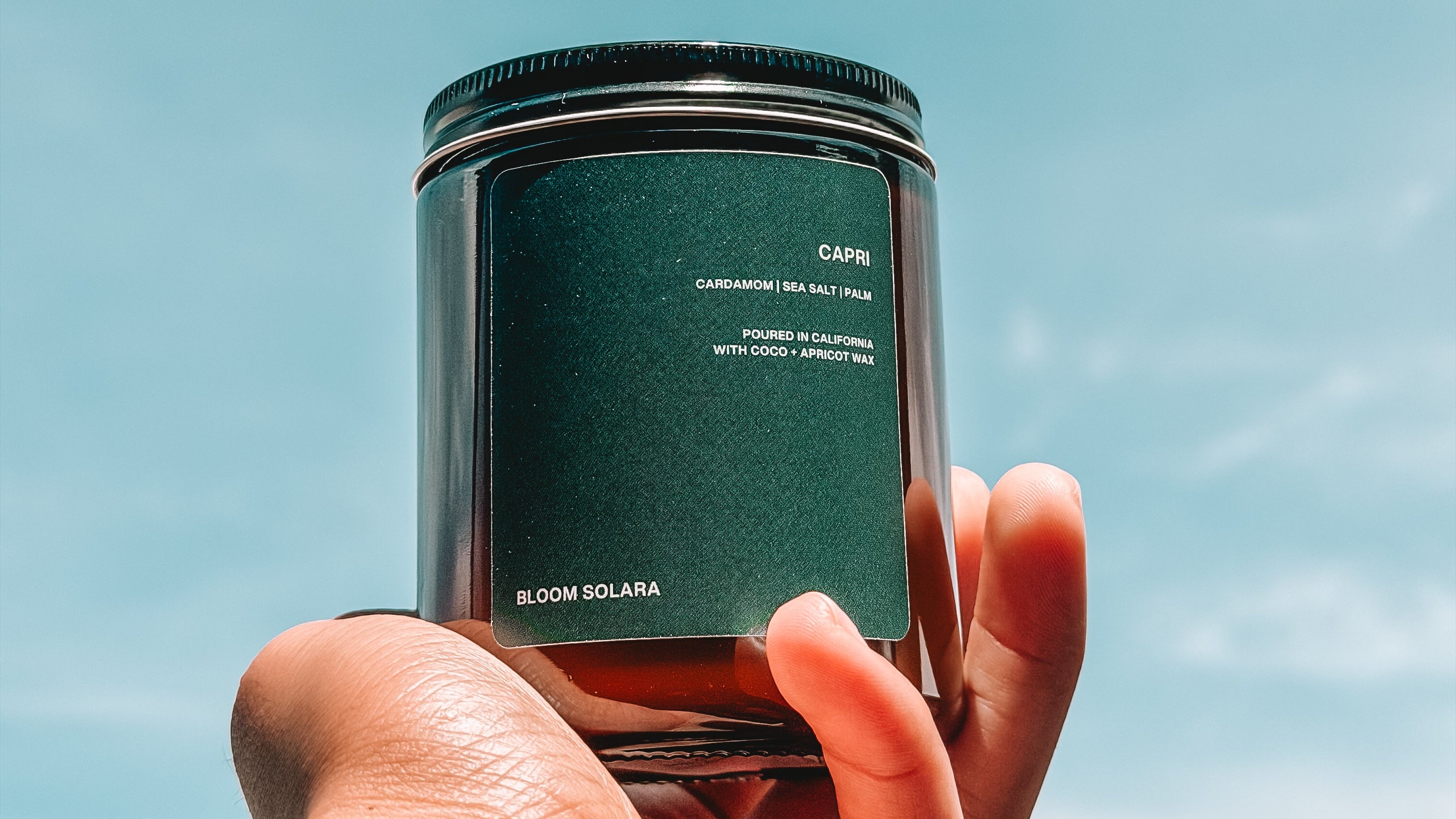 A hand holding a dark green glass jar candle against a bright blue sky background. The label reads 'COPK' and 'BLOOM SOLARA'.